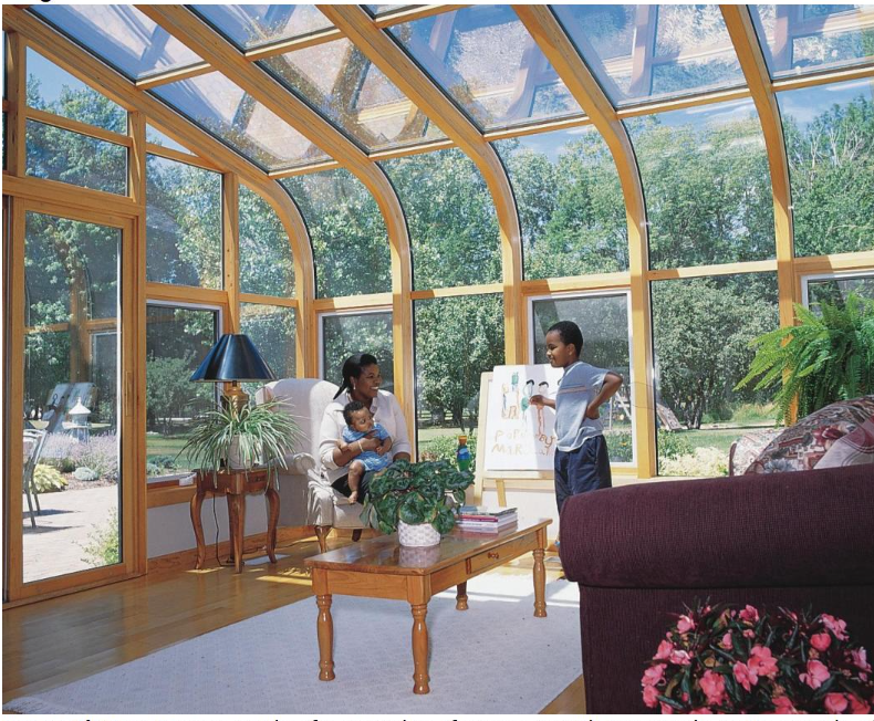 A scene inside a curved-roof sunroom with trees and a patio outside, of a woman sitting in a chair holding a child on her lap, while another older child stands next to her, pointing at something on an easel.
